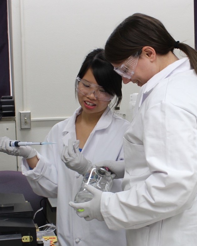 two CSN students in lab coats and safety goggles looking a research instrument